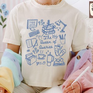 May include: Ivory Comfort Colors boxy tee shirt with a blue graphic design featuring a computer, books, and the text "The Queen of Queries." The shirt has short sleeves and a relaxed fit.
