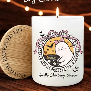 May include: A white soy candle with a wooden lid. The candle has a cartoon ghost design with the text "Smells Like Soup Season." The lid is engraved with "CUSTOM ENGRAVED LID." The candle is 10 oz.