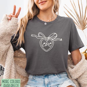 May include: Dark gray t-shirt featuring a white heart-shaped design with the letters "CPC" inside, adorned with a bow. The shirt is made of a soft material and is paired with denim shorts. The image also shows a beige cardigan.