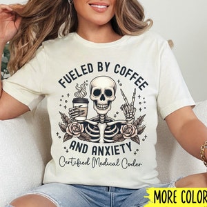 May include: A white t-shirt with a skeleton holding a coffee cup and a peace sign. The text on the shirt reads "Fueled by Coffee and Anxiety Certified Medical Coder".