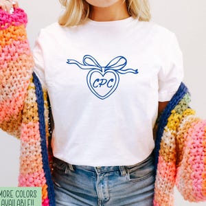 May include: White t-shirt with a blue heart-shaped design and the letters "CPC" inside. The shirt is paired with blue jeans and a colorful, striped cardigan. Text on the bottom left reads "MORE COLORS AVAILABLE!! WHITE BELLA CANVAS".