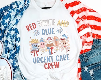 May include: White t-shirt with a red, white, and blue design featuring the text "Red White and Blue" with fireworks, a group of cartoon characters, and the text "Urgent Care Crew". The shirt is displayed on a model with a red, white, and blue patterned scarf and blue denim shorts.