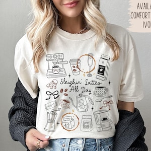 May include: Ivory-colored t-shirt with a coffee-themed graphic. The design includes coffee machines, cups, and the text "Sleighin' Lattes All Day." Additional text indicates more colors and comfort colors are available.