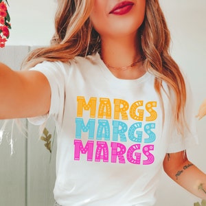 May include: White t-shirt with the word "MARGS" in three rows of colorful text. The top row is yellow, the middle row is blue, and the bottom row is pink. Each letter has a decorative design.