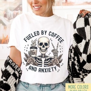 May include: White t-shirt with a skeleton holding a coffee cup and giving the peace sign. The text on the shirt reads "Fueled by Coffee and Anxiety".