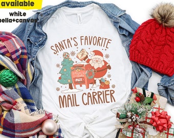 May include: White t-shirt with a red and green illustration of Santa Claus delivering mail. The text "Santa's Favorite Mail Carrier" is printed on the shirt.