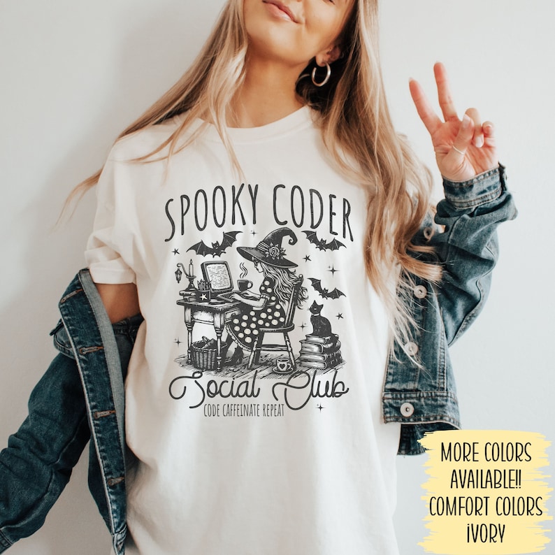 May include: White t-shirt with a black and white illustration of a witch coding at a computer. The text "Spooky Coder Social Club" is printed on the shirt. The text "Code Caffeinate Repeat" is printed below the illustration.