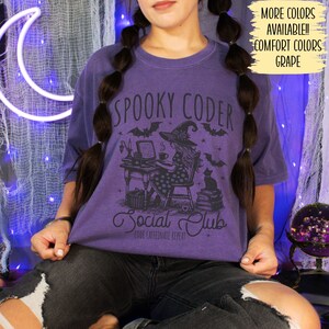 May include: Purple t-shirt with a graphic of a witch coding at a desk with a black cat and the text "Spooky Coder Social Club Code Caffeinate Repeat".