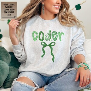May include: A grey sweatshirt with the word "coder" printed in green letters with a green bow underneath. The letters are decorated with a green shamrock pattern.