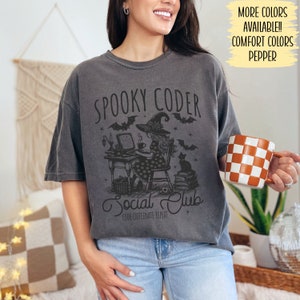 May include: A gray t-shirt with a black and white illustration of a witch coding at a computer. The text on the shirt reads "Spooky Coder Social Club Code Caffeinate Repeat".