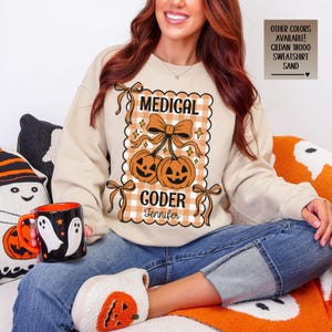 May include: Beige sweatshirt with a Halloween-themed design. The design features two orange jack-o'-lanterns, a bow, and the words "MEDICAL CODER Jennifer" in a decorative frame. The sweatshirt is a Gildan 18000 style.