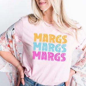 May include: A light pink t-shirt with the word "MARGS" printed three times in stacked rows. Each word is in a different color: yellow, blue, and pink. The letters have decorative patterns.