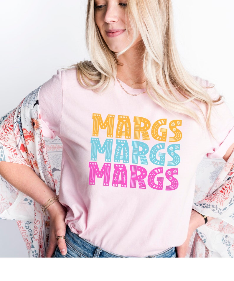 May include: A light pink t-shirt with the word "MARGS" printed three times in stacked rows. Each word is in a different color: yellow, blue, and pink. The letters have decorative patterns.