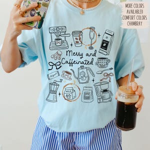 May include: Light blue t-shirt with a black and white coffee-themed graphic. The design includes coffee-making equipment, coffee beans, and the text "Merry and Caffeinated." Additional text indicates more colors are available.