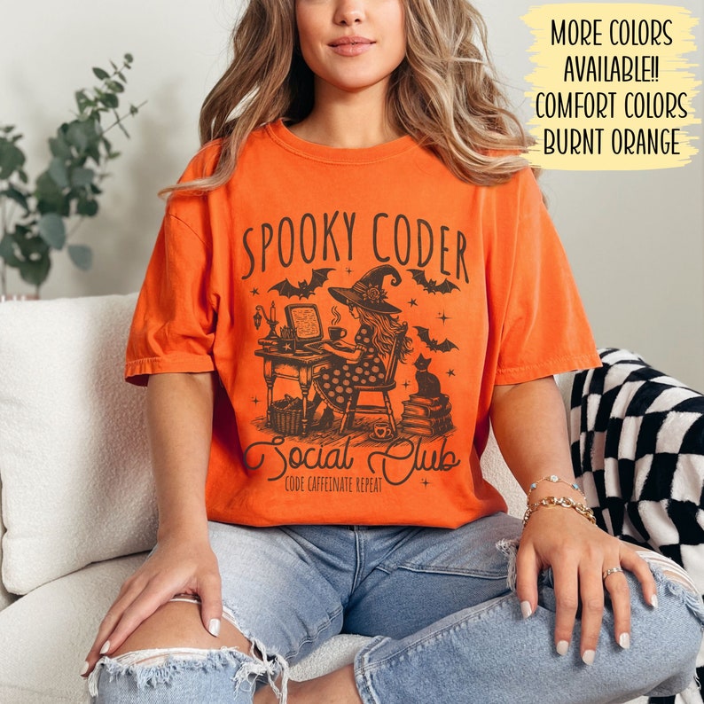 May include: A burnt orange t-shirt with a graphic of a witch coding at a computer with a black cat and the text "Spooky Coder Social Club Code Caffeinate Repeat".