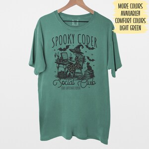 May include: A light green t-shirt with a black and white illustration of a witch coding on a computer. The text "Spooky Coder Social Club" is printed on the shirt. The text "Code Caffeinate Repeat" is printed below the illustration.