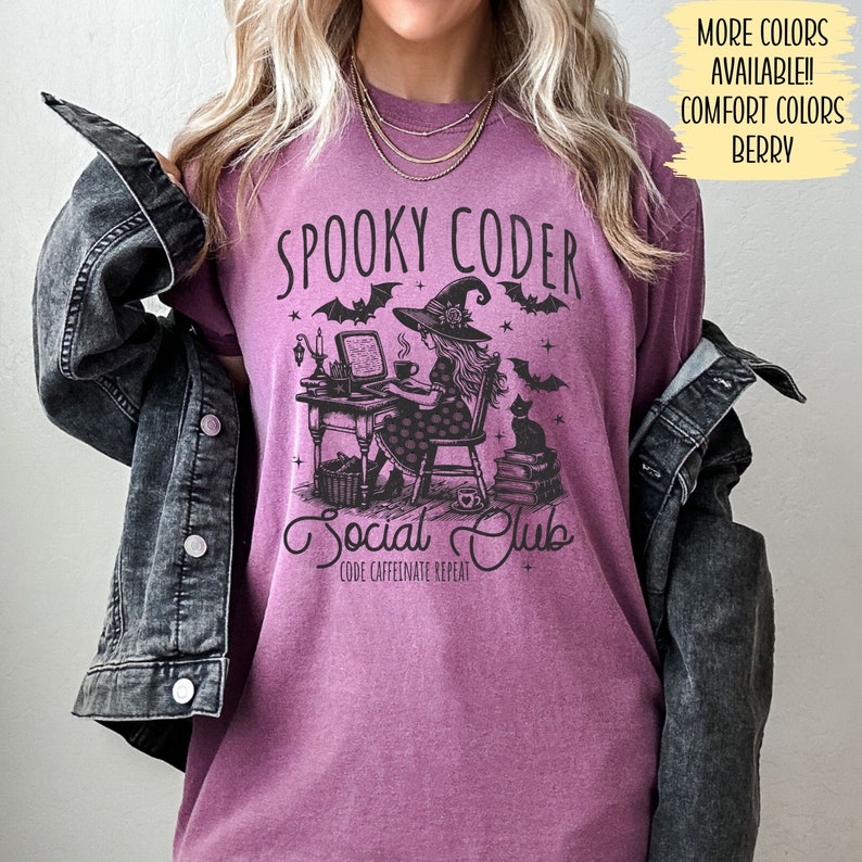 May include: A purple t-shirt with a black graphic of a witch coding on a computer. The text on the shirt reads "Spooky Coder Social Club Code Caffeinate Repeat".