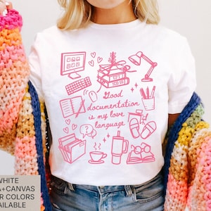 May include: White Bella+Canvas t-shirt with a pink graphic design featuring a computer, books, a lamp, and the text "Good documentation is my love language." The shirt is paired with a colorful knitted sweater.