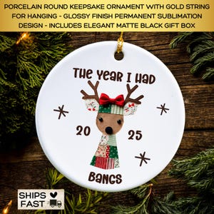 May include: A white porcelain round keepsake ornament with a gold string for hanging. The ornament features a reindeer design with the text "The year I had 2025 bangs". The reindeer has brown antlers, a red bow, and a patchwork design.