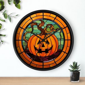 Puede incluir: Un reloj con marco negro con un diseño de vitral de una calabaza de Halloween con una cara sonriente. La esfera del reloj está rodeada por un borde decorativo naranja y marrón.