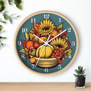 May include: A round clock with a blue face and white numbers. The clock features a fall harvest theme with sunflowers, pumpkins, and other fruits and vegetables in a basket.