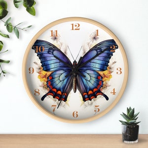 Könnte beinhalten: Eine weiße Uhr mit einem blauen Schmetterlingsdesign und schwarzen Zeigern. Das Zifferblatt der Uhr hat römische Ziffern für die Stunden und arabische Ziffern für die Minuten. Der Schmetterling befindet sich in der Mitte des Zifferblatts und hat orange und rote Akzente.