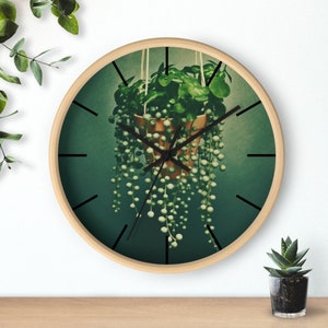 Könnte beinhalten: Eine runde Uhr mit Holzrahmen, schwarzen Zeigern und grünem Hintergrund. Eine Topfpflanze mit grünen Blättern und langen, perlenartigen Ranken hängt an der Oberseite der Uhr.