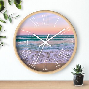 May include: A round wooden clock with a beach scene and Roman numeral hour markers. The clock face shows a sunset over the ocean with waves crashing on the shore.
