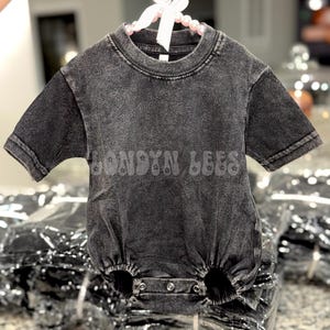 May include: A charcoal gray baby romper with short sleeves and a round neckline. The romper features the text "LONDYN LEES" in a faded, textured font across the front. It is hanging on a white pearl beaded hanger with a white bow.