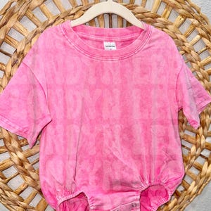 May include: A bright pink baby romper with a tie-dye effect, hanging on a wooden hanger. The romper has short sleeves and snaps at the bottom. The romper is displayed against a woven wicker background.