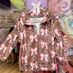 May include: A brown and black leopard print pullover with pink bow accents. The top has a white zipper and the word "LONDYN" is printed on the collar. The long-sleeved top is hanging on a pink pearl beaded hanger.