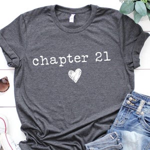 May include: Dark gray t-shirt with the text "chapter 21" and a heart graphic. The shirt is paired with blue jeans, white sneakers, and sunglasses. A sprig of greenery is also visible.