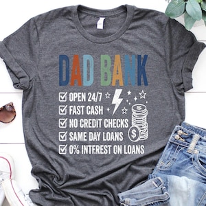 May include: A dark gray t-shirt with the text "DAD BANK" in blue and orange. The shirt features a list of services, including "OPEN 24/7" and "0% INTEREST ON LOANS". Sunglasses, jeans, and sneakers are also in the image.