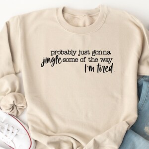 Puede incluir: Una sudadera beige con el texto "probably just gonna jingle some of the way I'm tired" en escritura negra. La sudadera se combina con vaqueros azul claro y zapatillas blancas.