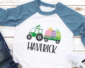 May include: A white and blue baseball tee shirt with a green tractor carrying colorful Easter eggs. The shirt has the name "Maverick" printed on it.