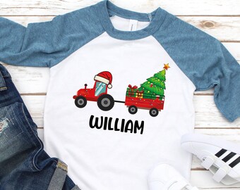 May include: A white and blue baseball tee shirt with a red tractor pulling a wagon with a Christmas tree. The text "William" is printed below the tractor.