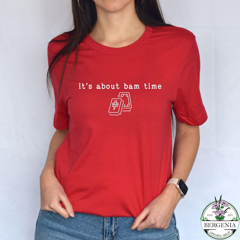 Puede incluir: Camiseta roja con la frase "it's about bam time" en blanco y un gr&aacute;fico de dos fichas de Mahjong. La camiseta tiene cuello redondo y mangas cortas. La tela parece suave y c&oacute;moda.