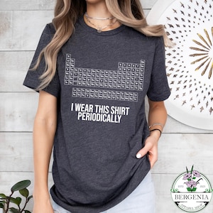 May include: Dark gray t-shirt featuring a white periodic table design. The shirt includes the text "I wear this shirt periodically". The shirt is a short-sleeved crew neck.