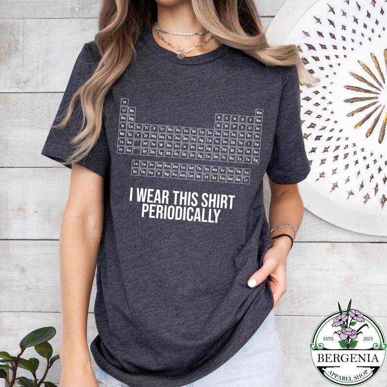 May include: Dark gray t-shirt featuring a white periodic table design. The shirt includes the text "I wear this shirt periodically". The shirt is a short-sleeved crew neck.