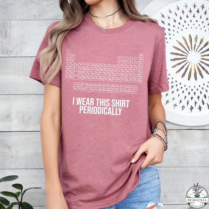 May include: A mauve t-shirt featuring a white periodic table design and the text "I wear this shirt periodically." The shirt is a short-sleeved crew neck style.