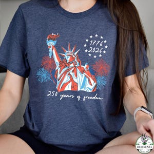 May include: Navy blue t-shirt featuring a graphic of the Statue of Liberty in red, white, and blue, holding a torch. The shirt includes the text "1776 2026" and "250 years of freedom" with fireworks in the background.