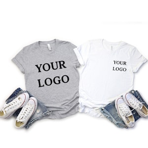 Puede incluir: Dos camisetas, una gris y otra blanca, con el texto "YOUR LOGO" impreso en ellas. Las camisetas est&aacute;n dispuestas sobre un fondo blanco con un par de pantalones cortos de mezclilla azul y zapatillas blancas.