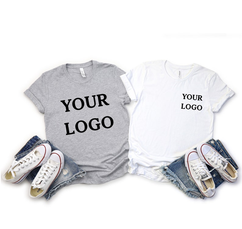 Puede incluir: Dos camisetas, una gris y otra blanca, con el texto "YOUR LOGO" impreso en ellas. Las camisetas est&aacute;n dispuestas sobre un fondo blanco con un par de pantalones cortos de mezclilla azul y zapatillas blancas.