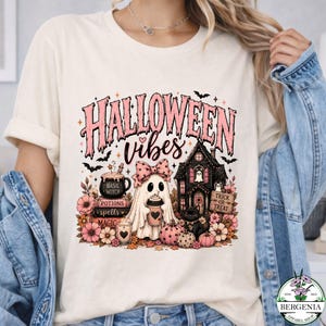 May include: A cream-colored t-shirt featuring a graphic with the text "HALLOWEEN vibes". The design includes a ghost wearing a pink bow, a haunted house, a black cat, a stack of books labeled "BASIC WITCH", "POTIONS", "spells", and "MAGIC", and pumpkins.