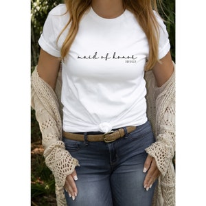 May include: White t-shirt with the text "maid of honour" in black cursive writing and the word "obviously" in smaller print below. The shirt is being worn by a person with a brown belt and blue jeans.