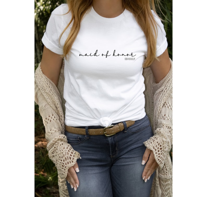 May include: White t-shirt with the text "maid of honour" in black cursive writing and the word "obviously" in smaller print below. The shirt is being worn by a person with a brown belt and blue jeans.