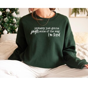 Puede incluir: Sudadera verde oscuro con el texto blanco: "probably just gonna jingle some of the way I'm tired". La sudadera de cuello redondo tiene mangas largas. Un &aacute;rbol de Navidad y un sof&aacute; blanco son visibles en el fondo.