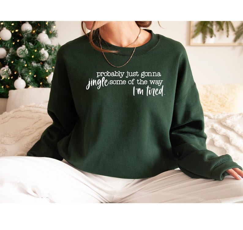 Puede incluir: Sudadera verde oscuro con el texto blanco: "probably just gonna jingle some of the way I'm tired". La sudadera de cuello redondo tiene mangas largas. Un &aacute;rbol de Navidad y un sof&aacute; blanco son visibles en el fondo.