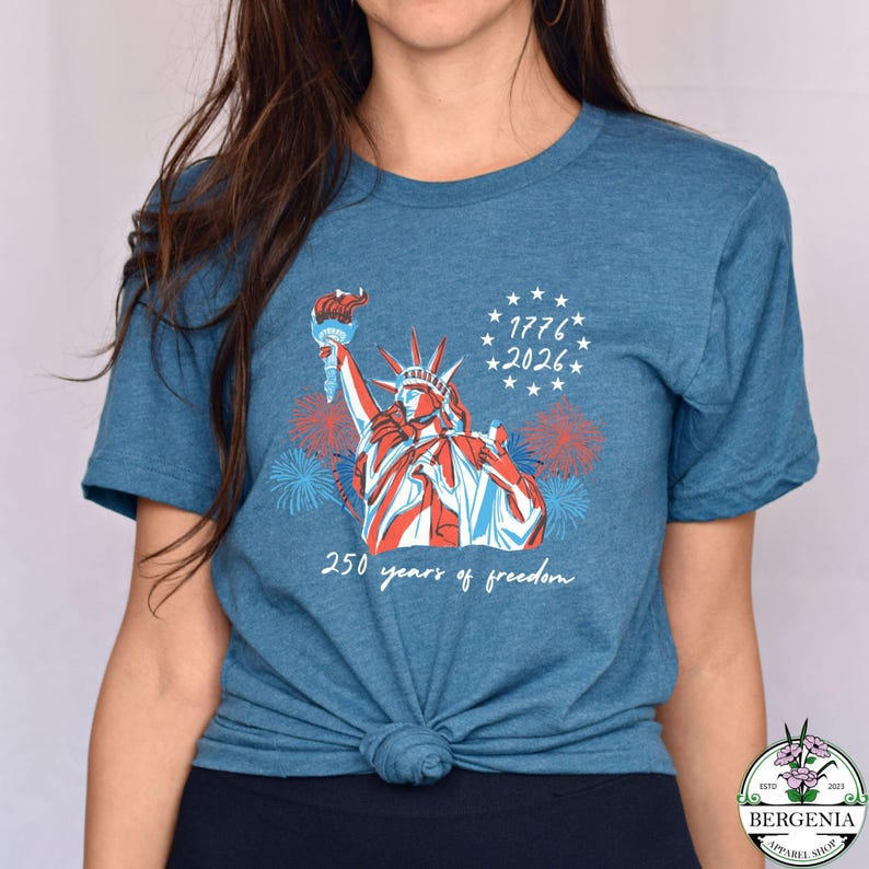 May include: A blue t-shirt featuring a graphic of the Statue of Liberty in red, white, and blue, holding a torch. The shirt includes the text "1776 * 2026" and "250 years of freedom."