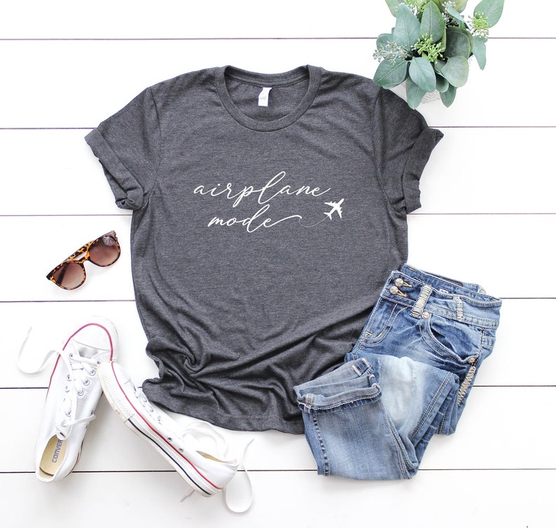 May include: A dark grey t-shirt with the text "airplane mode" in white cursive writing and two small airplane icons. The shirt is laid out on a white wooden surface with a pair of sunglasses, white trainers, and blue jeans.